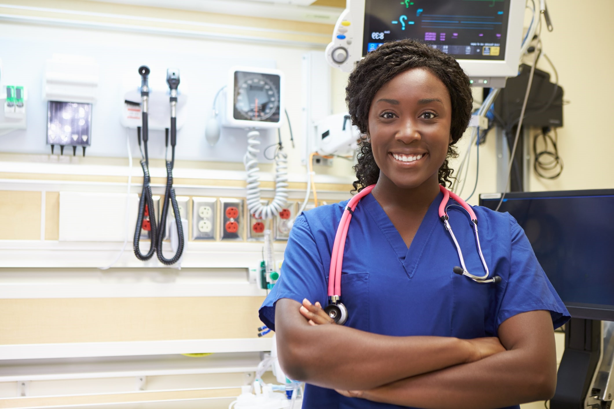 ER travel nurse in blue scrubs stands smiling in front ER tools 